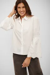 Camisa blanca de corte clásico con cuello camisero y cierre frontal de botones. Destaca por sus mangas largas con detalle de frunce a la altura del codo y terminación acampanada con bordado inglés.