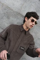 Camisa de corte boxy con estampado de cuadros pequeños en tonos oscuros. Presenta cuello camisero en color negro, cierre frontal con botones, canesú en la parte superior y un bolsillo en el pecho con etiqueta de la marca Kapsul Studio.