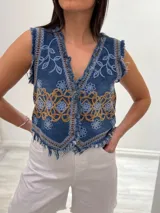 Chaleco corto en denim azul con detalle desflecado y bordado artesanal en tonos celeste y dorado. Estilo boho chic con escote en V, ideal para combinar con prendas básicas y elevar tu look.
