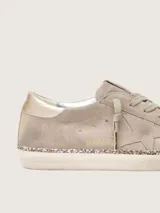 Zapatillas Golden Goose modelo Super-Star en gamuza color topo. Presentan diseño de aspecto desgastado, con la clásica estrella lateral en el mismo tono, contrafuerte metalizado en color bronce, cordones tono sobre tono y detalle de glitter champagne en el canto de la suela.