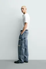 Pantalón de jean estilo carpintero con corte relaxed fit, tiro medio y pierna recta. Presenta un lavado azul medio con efecto desgastado y roturas en las rodillas. Incluye bolsillos laterales tipo plastrón, tira multifunción en la pierna, cierre frontal con cremallera y botón.