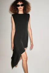 Vestido corto negro sin mangas con hombreras, cuello redondo y bajo asimétrico con una caída lateral más larga. Cuenta con cierre en la espalda con abertura y botón.