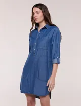 Vestido camisero blanco de rayón, con cuello con solapa, escote en V y pechera de abertura frontal con botones. Presenta un bolsillo delantero, bolsillos laterales aplicados y mangas largas con puño de largo regulable con presilla y botón. Cuenta con calce holgado y caída ligera, con ruedo irregular más largo en la parte trasera.
