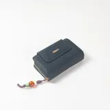 Billetera negra de textura granulada con cierre de cremallera dorada y solapa frontal con broche magnético. Incluye aplique metálico con el logo de la marca en letras doradas y un cordón decorativo colgante con cuentas de colores.