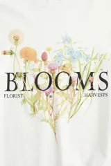 Remera de algodón color blanco crudo, corte oversized y cuello redondo. Presenta un estampado central de flores silvestres con la palabra "BLOOMS" en tipografía serif negra, y las palabras "FLORIST" y "HARVESTS" a los lados.