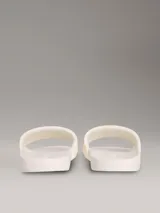 Ojotas tipo slide de color blanco hueso, con tira ancha acolchada y monograma de CK en relieve. La suela es de espuma.