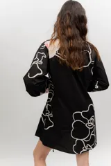 Vestido corto de popelina color negro con estampado de líneas blancas y un diseño floral destacado en el pecho. Cuenta con escote en V, cuello clásico, mangas abombadas y detalles de cordones laterales con efecto corsé.
