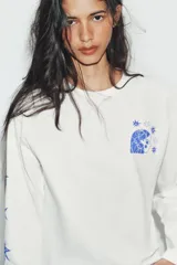 Remera de manga larga y corte oversize, color blanco, con cuello redondo. Presenta un estampado gráfico en color azul en el pecho y a lo largo de la manga derecha.