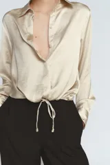 Camisa de cuello solapa y manga larga con puños anchos, confeccionada en tejido satinado color beige. Presenta cierre frontal con botones ocultos y bajo ajustable mediante cordones del mismo tejido.
