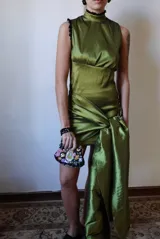 Vestido corto de tafeta color verde oliva, de cuello alto y sin mangas, con drapeados en el cuerpo y un gran nudo lateral con cola larga.