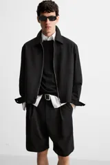 Bermuda de corte relajado color negro, con pliegues frontales, cierre de cremallera y botón, bolsillos laterales y bolsillos traseros.