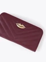 Maxi billetera de cuero vacuno con diseño matelassé en forma de V. Presenta un aplique metálico dorado en forma de labios en el frente y cierre perimetral con tirador de cuero.