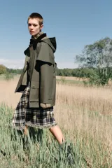 Trench de estilo encerado color verde militar con cuello de solapa en pana marrón, capucha extraíble y capa superior. Presenta cierre frontal con cremallera y botones a presión ocultos, bolsillos delanteros de plastrón con solapa y mangas largas con trabillas.