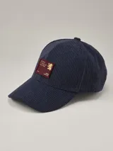 Gorra de béisbol azul marino confeccionada en pana, con parche rectangular bordó en el frente que presenta un diseño gráfico y texto en color dorado.