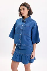 Sobrecamisa de jean de manga corta y corte oversize en color azul medio, con bolsillos frontales con solapa y cierre de botones metálicos a presión.