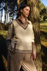 Sweater de punto estilo preppy en color crudo con detalles en marrón. Presenta cuello en V con ribete a rayas, tejido con textura de ochos y cordones laterales decorativos. Posee puños y ruedo acanalados con diseño a rayas.