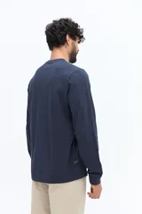 Remera de manga larga color blanco roto o gris muy claro, con escote redondo y un pequeño logo circular estampado en el pecho.