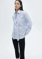 Camisa de popelín de algodón en color celeste claro, con cuello camisero, cierre frontal de botones y detalle de lazada tipo corbata integrada al cuello. Presenta un bolsillo de parche en el pecho, corte recto y mangas largas con puños abotonados.