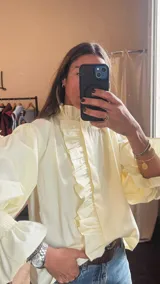 Camisa marrón de cuello alto con volados, mangas abullonadas y puños elásticos fruncidos. Cuenta con una tira de volados en la parte frontal.