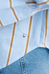 Camisa de corte oversize con estampado de rayas verticales en tonos azul y beige. Presenta cuello camisero clásico, cierre frontal con botones, bolsillo en el pecho y mangas largas con puños rectos.