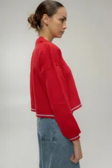 Cardigan de punto color rojo, con cuello redondo, cierre frontal de botones y mangas largas. Presenta un detalle bordado de una concha marina en el pecho y terminaciones en contraste de color celeste en el ruedo, puños y bolsillo frontal.