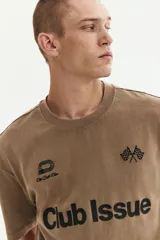 Remera de algodón de punto pesado con efecto lavado, color beige/tostado. Presenta un corte boxy (holgado) con cuello redondo ribeteado y bajo recto. Estampado frontal en negro con texto "Club Issue" y gráficos de banderas a cuadros.