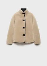 Chaqueta de efecto pelo en color beige, con diseño oversize y corte recto. Presenta cuello alto, cierre frontal con botones y detalles de ribetes en cuero sintético negro.