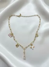 Collar dorado con cadena de eslabones rectangulares y varios dijes colgantes: elefante rosa, corazón calado, avión blanco y naranja, moneda dorada y Torre Eiffel rosa.