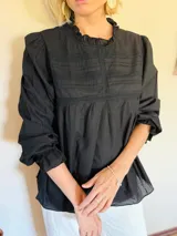 Blusa negra de manga larga con detalles de encaje y frunces en el pecho. Presenta cuello redondo con volados y puños con elástico y volado.
