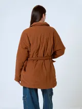 Campera de pana color marrón, con diseño acolchado, cuello camisero, cierre frontal oculto y cinturón ajustable con hebilla a tono. Presenta bolsillos frontales con solapa y mangas largas de corte holgado.