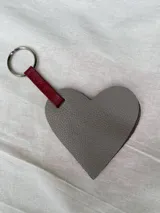 Llavero con forma de corazón de cuero gris con tira de cuero rojo y aro metálico.