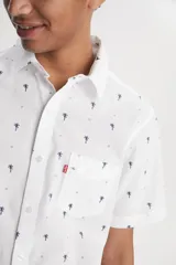 Camisa de manga corta con cuello clásico y cierre frontal de botones. Presenta un estampado de pequeñas palmeras oscuras sobre fondo blanco y cuenta con un bolsillo frontal en el pecho con etiqueta roja distintiva de la marca.