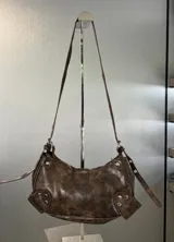 Cartera tipo hobo de cuero sintético con efecto desgastado, color marrón. Presenta una hebilla central decorativa, detalles de tachas metálicas en los laterales y correa ajustable.