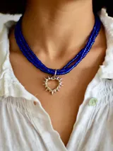 Collar de tres vueltas compuesto por pequeñas cuentas de vidrio en color azul intenso, con un dije central en forma de corazón con diseño de rayos en metal plateado.