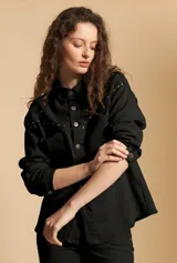 Sobrecamisa negra de estilo estructurado con cierre frontal de botones metálicos, cuello clásico y detalle de tachas decorativas en la zona de los hombros y el pecho. Presenta mangas largas con puños abotonados.