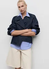 Abrigo corto estilo chaqueta de jean, confeccionado en algodón denim azul oscuro. Presenta cuello con solapas, cierre frontal, trabillas en los hombros y mangas largas con trabillas ajustables.