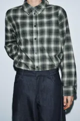 Camisa de corte oversize confeccionada en algodón, con estampado de cuadros en tonos verde, negro y blanco. Presenta cuello solapa, cierre frontal con botones, manga larga con puños y bajo asimétrico.