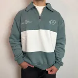 Buzo de algodón con cuello polo y cierre frontal parcial. Presenta un diseño de bloques de color con una franja horizontal blanca central sobre una base de color verde azulado. Incluye detalles bordados en el pecho y puños elásticos.