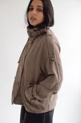 Chaqueta de corte estructurado con estampado cuadrille en tonos marrones y beige. Presenta cuello alto con tira ajustable, cierre frontal con botones, charreteras en los hombros y puños con detalle de hebilla.