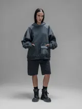 Hoodie oversize de color gris oscuro, confeccionado en frisa de algodón con recortes en textiles técnicos. Presenta capucha forrada, bolsillo frontal tipo canguro y un bolsillo adicional con solapa en la parte inferior.