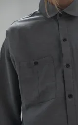 Camisa de manga larga en tejido twill color gris melange, con corte oversize. Presenta cuello clásico, cierre frontal con botones y tres bolsillos delanteros, uno de ellos oculto.