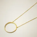 Collar de cadena fina con dije circular, elaborado en plata 925 con baño de oro. El dije presenta un diseño minimalista con incrustaciones de circonias en dos secciones opuestas del círculo.