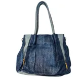 Cartera tipo tote de diseño estructurado, combinando paneles de tela denim clara con secciones centrales de tejido texturizado en azul oscuro. Presenta cierres metálicos decorativos en los laterales y doble asa de hombro.