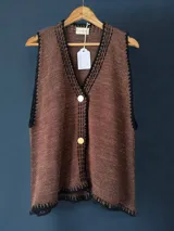 Chaleco tejido color tostado con destellos de lurex, escote en V y cierre frontal con botones. Presenta detalles de bordados y terminaciones en crochet en color crudo.