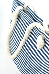 Bolso tipo tote de playa, con diseño de rayas horizontales en color gris y base blanca. Cuenta con asas de soga gruesa en color crudo con nudos decorativos.