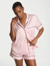 Pijama corto de satén color rosado con detalles de ribetes negros, compuesto por una camisa de manga corta abotonada con bolsillo en el pecho y el logotipo bordado en el cuello, y un short con cintura elástica.
