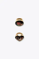 Pack de dos anillos metálicos dorados con detalles en resina negra y diseños florales. Uno de los anillos es ovalado con flores rojas y fucsias, y el otro tiene forma de corazón con un pájaro y flores.