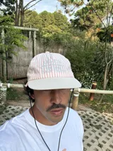 Gorro de visera curva con diseño a rayas verticales en tonos rosado pálido y blanco. La corona tiene un bordado en azul claro con texto. La visera es de color beige claro. Incluye un cordón negro ajustable para colgar del cuello.