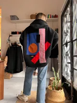 Campera de textura tipo corderito en color negro, con diseño de collage geométrico en la espalda que incluye parches en azul, naranja, marrón y franjas rojas y blancas. Posee capucha y mangas largas.