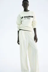 Buzo de tejido de punto color blanco roto, con cuello redondo, hombros caídos y puños y bajo acanalados. Presenta un estampado frontal en negro con el texto "LE MAISON de Paris".
